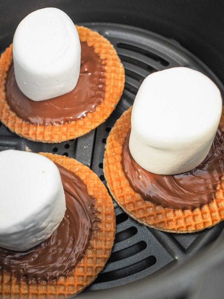 A look inside an air fryer basket with three stroopwaffels spread with Nutella and a large marshmallow before air frying.