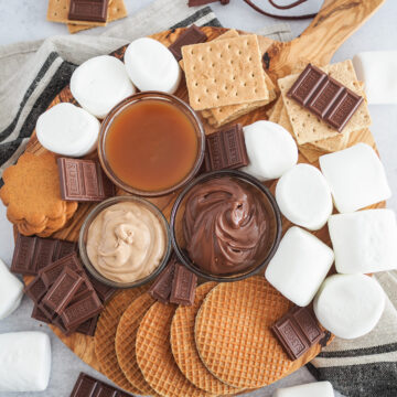 Round wood board filled with ingredients to make air fryer s'mores including marshmallows, graham crackers, hershy's chocolate, stroopwaffels, cookies, nuttella, caramel, and cinnamon spread.