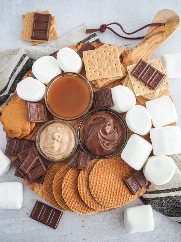 Round wood board filled with ingredients to make air fryer s'mores including marshmallows, graham crackers, hershy's chocolate, stroopwaffels, cookies, nuttella, caramel, and cinnamon spread.