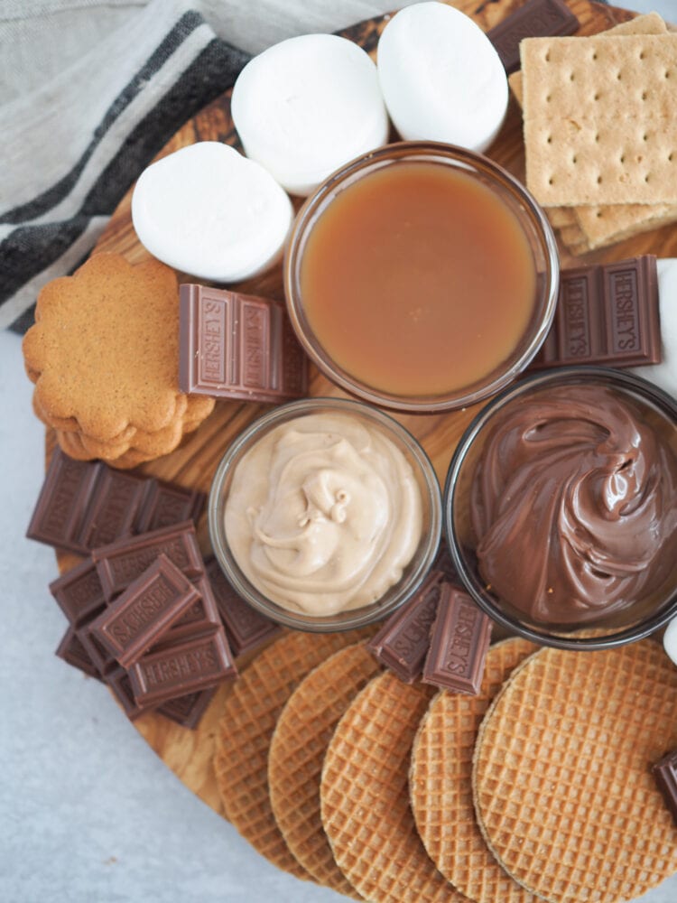 Round wood board filled with ingredients to make air fryer s'mores including marshmallows, graham crackers, hershy's chocolate, stroopwaffels, cookies, nuttella, caramel, and cinnamon spread.