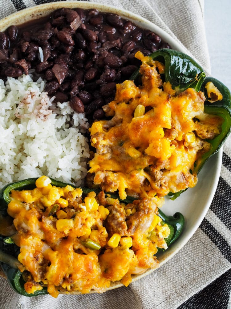 Two stuffed poblano peppers on a dinner plate with white rice and black beans.