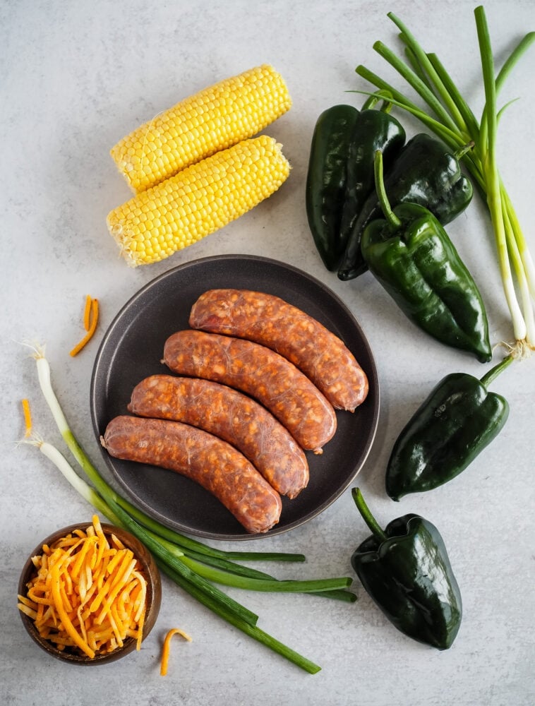 Overhead look at the ingredients to make chorizo stuffed poblano peppers including corn on the cob, fresh chorizo sausage, green onions, cheddar cheese, and poblano peppers.