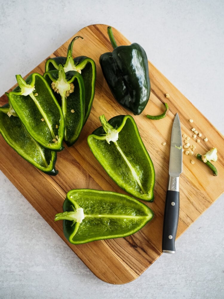 Wood cutting board with a knife and dark green poblano peppers cut in half.
