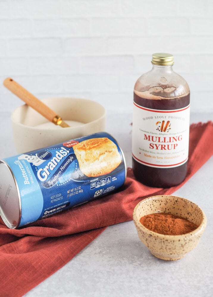 Ingredients to make air fried apple cider donuts including a can of buttermilk biscuit dough, mulling syrup, cinnamon, and a bowl of sugar all placed beside an orange autumnal napkin.