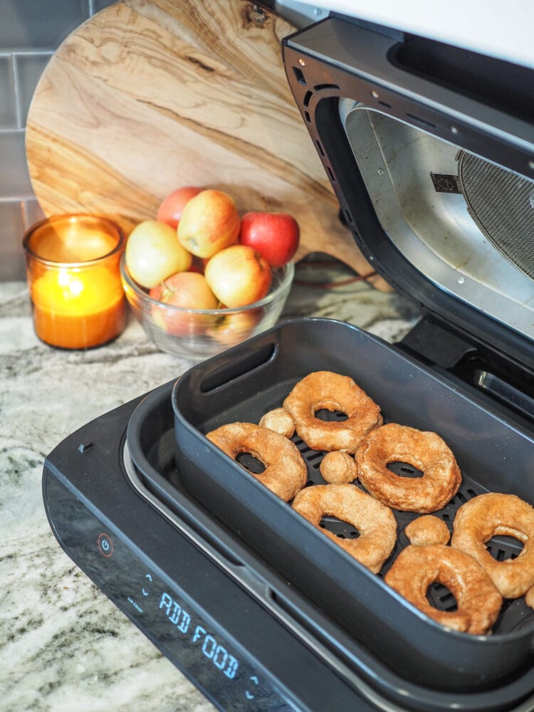 Raw donuts placed in Ninja 6-in-1 Air Fryer Grill before air frying, There's a bowl of apples and orange candle in the background.