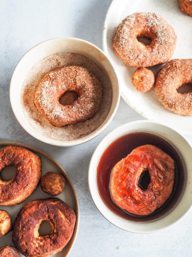 Four plates to assemble apple cider air fried donuts including golden air fried donuts, donut in a bowl with mulling spices syrup, donut in a bowl of cinnamon sugar, and plate with fully syrup and cinnamon sugar dipped donuts.