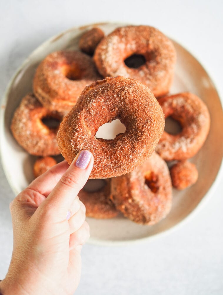 Hand with purple glitter fingernail polish holding a single air fried donut over a plate of donuts.