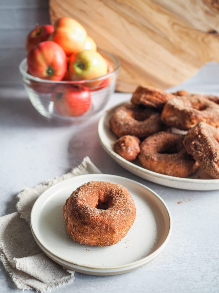 Single air fried donut on a plate with a bowl of red apples and a plate of donuts and donut holes in the background.