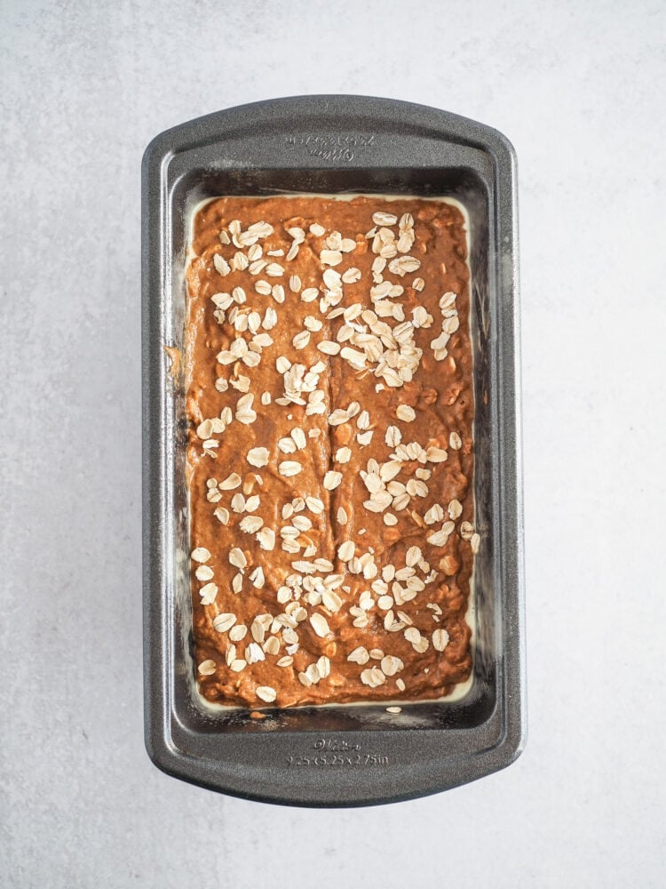 Overhead look at a bread loaf pan filled with Guinness Irish brown bread dough before baking. It has a small slit in the center drawn by a spatula to let the bread vent a it bakes. And it is sprinkled with oats on top.