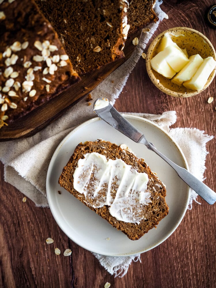 Overhead look at a slice of Irish brown bread on a ceramic plate, butter spread across the surface of the bread. There is a loaf of brown bread and a dish of butter on the dark wood table as well.
