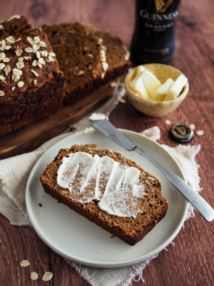 A loaf of Irish brown bread on a dark wood table. There's a slice of brown bread with butter on a ceramic plate, a natural cloth napkin, dish of butter, and bottle of Guinness.