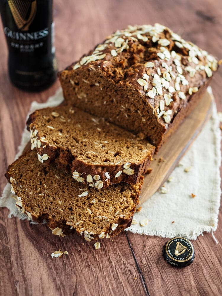 Loaf of Irish brown bread sliced on a wood cutting board, placed on a towel resting on a dark wood table. There's a bottle of Guinness in the background.