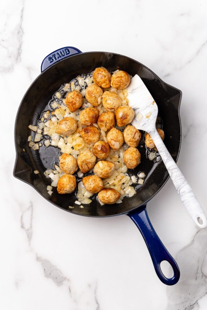 Blue Staub skillet with meatballs and diced onion cooking in butter.