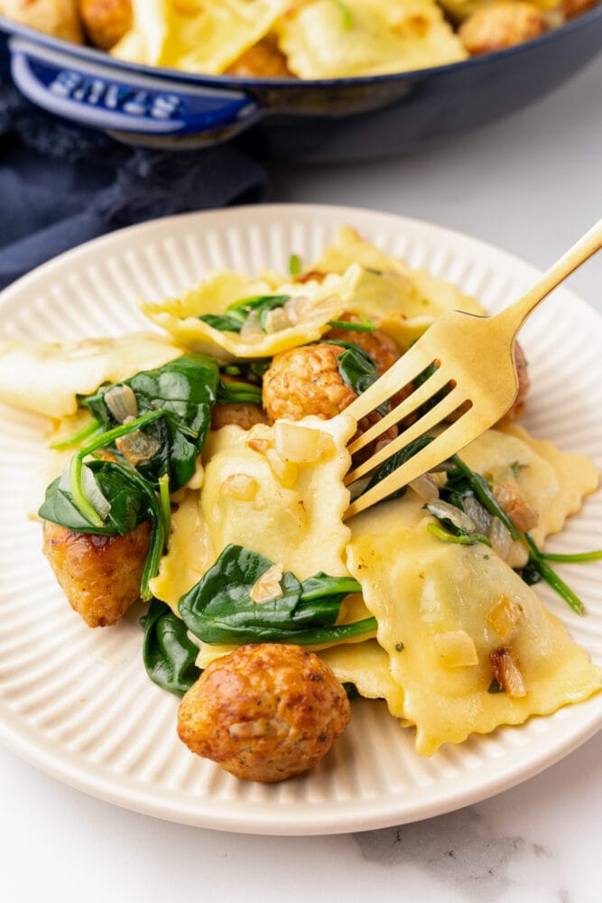 Small plate of ravioli and meatballs with spinach. There's a gold fork getting ready to take a piece, an a blue skillet of ravioli in the background.