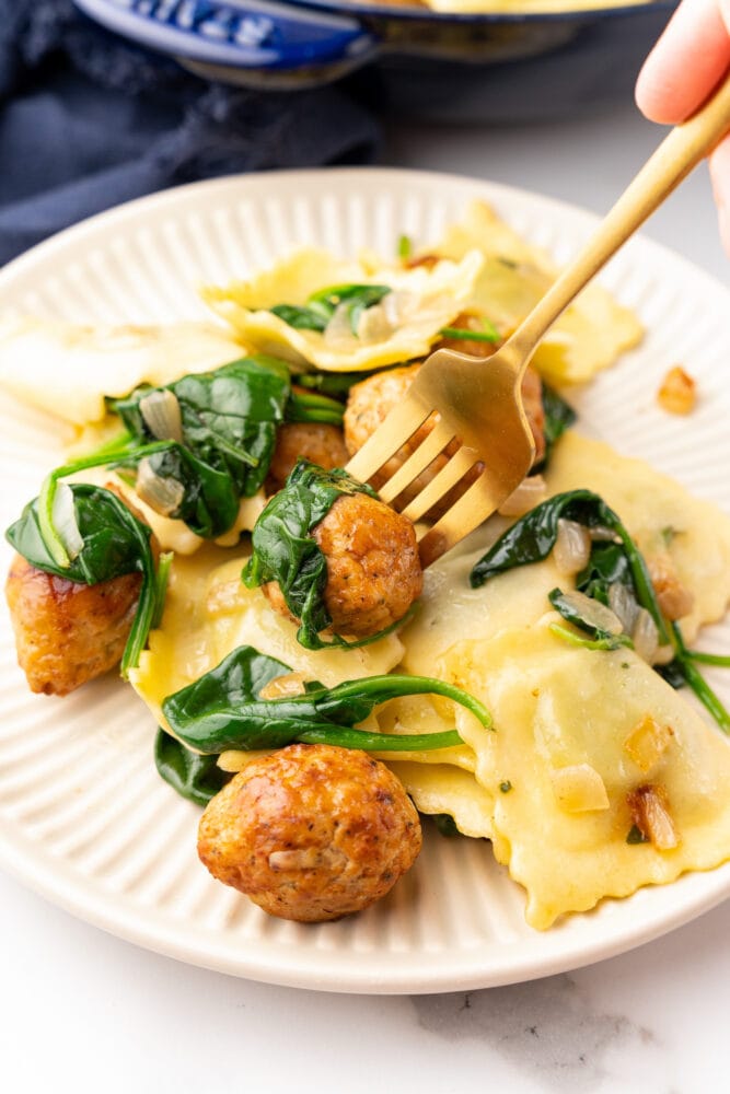 Small plate of ravioli and meatballs with spinach. There's a gold fork getting ready to take a piece, an a blue skillet of ravioli in the background.