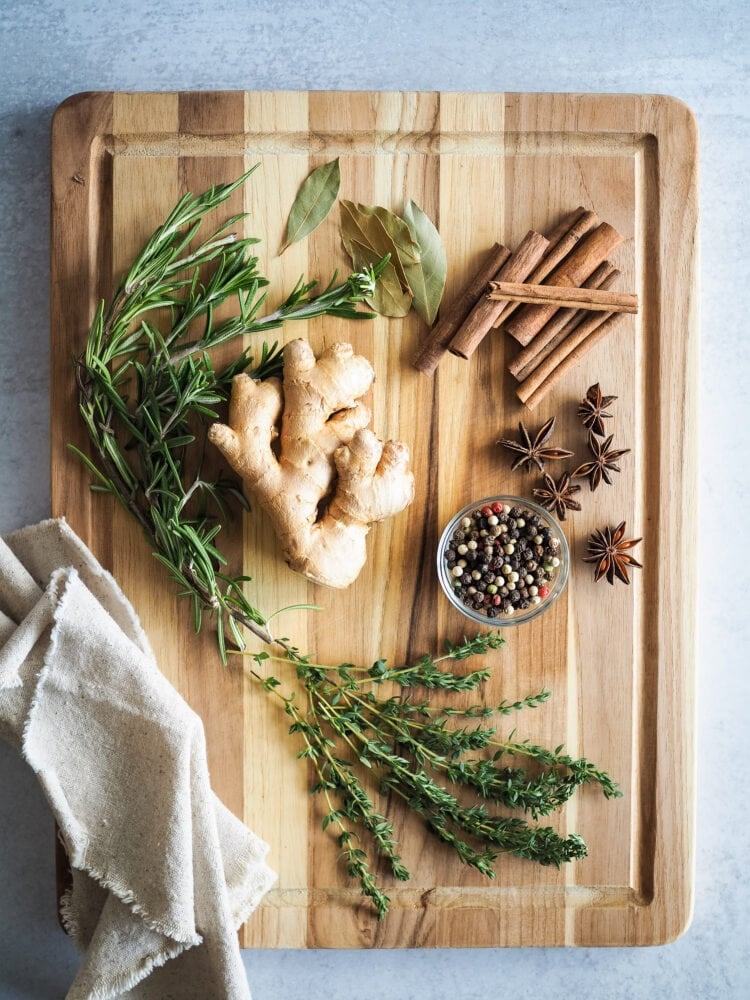 wood cutting board with variety of ingredients to make a simmer pot including ginger, rosemary, thyme, bay leaves, cinnamon sticks, peppercorns, and star anise