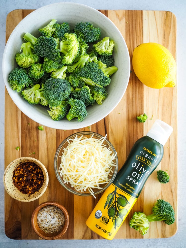 Overhead view of air fried broccoli ingredients including bowl of small pieces of broccoli florets, lemon, shredded parmesan cheese, red pepper flakes, salt, pepper, and olive oil spray.