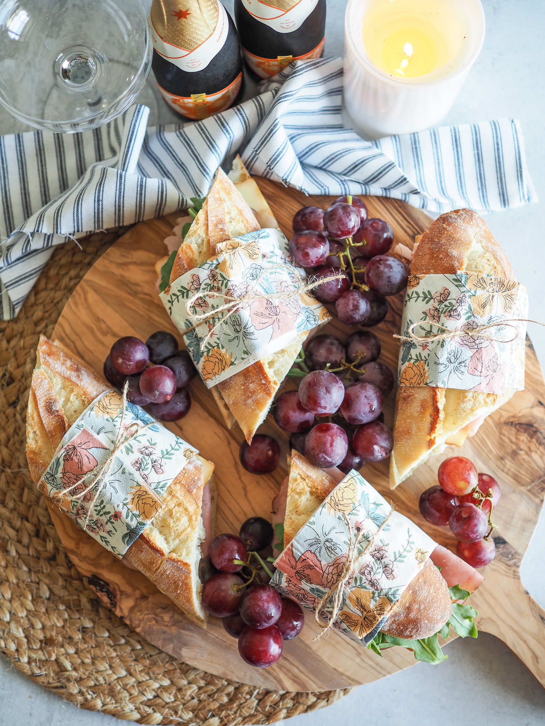 Four baguette sandwiches wrapped in napkins and twine. They're placed on a wood board, surrounded by grapes. Theres's champagne and a candle off to the side.
