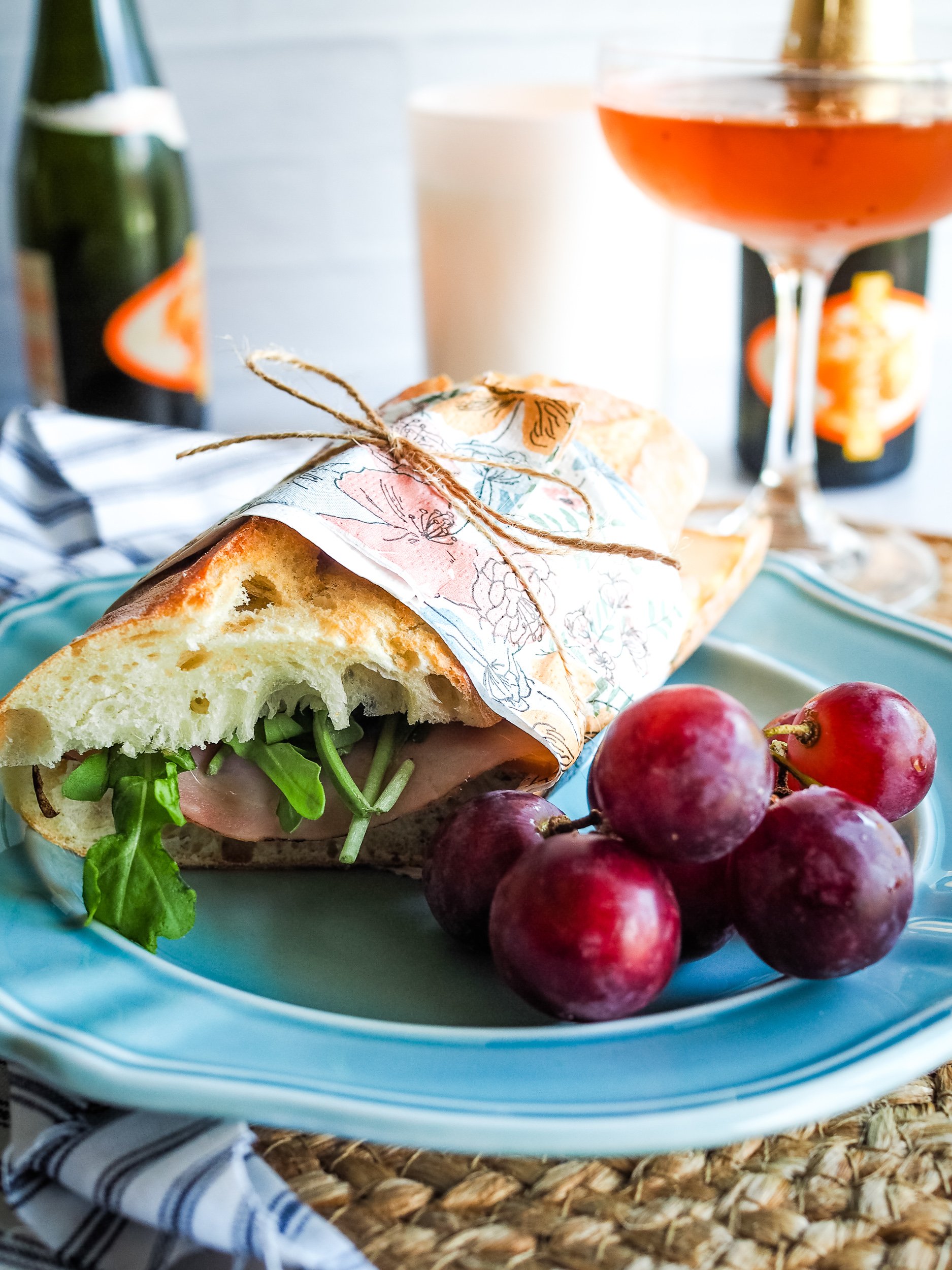 Side view of baguette sandwich filled with ham and arugula, wraped in floral napkin, tied with twine, and set onto a blue plate with grapes. There's a candle and rose champagne in the background.