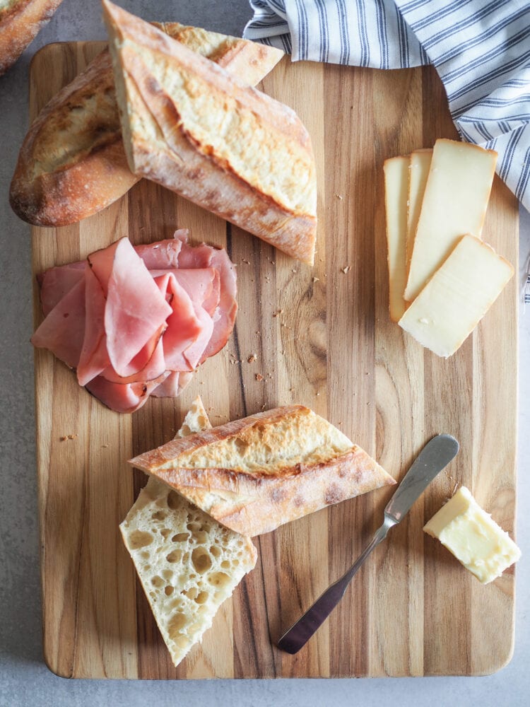 Baguette slice on a cutting board with slices of ham, slices of cheese, butter, and a spreader off to the side.