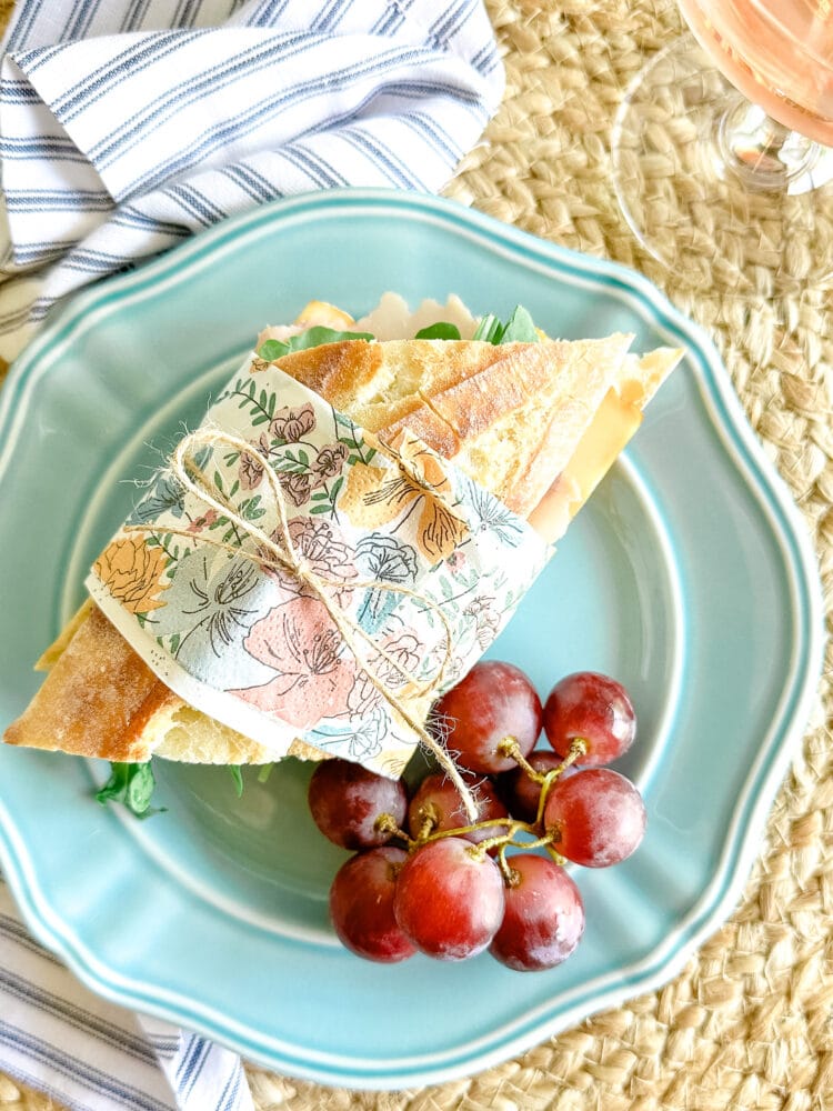 Baguette sandwich wrapped in a floral napkin and twine on a blue plate. There are grapes accompanying the sandwich and rose in a glass off to the side as well as a blue and white striped napkin.