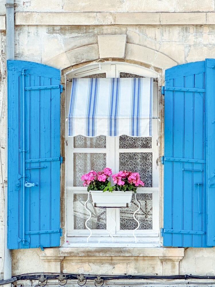 A beautiful window with teal blue shutters, a blue and white stripped awning, pink flowers, and lace curtains in Arles, France.