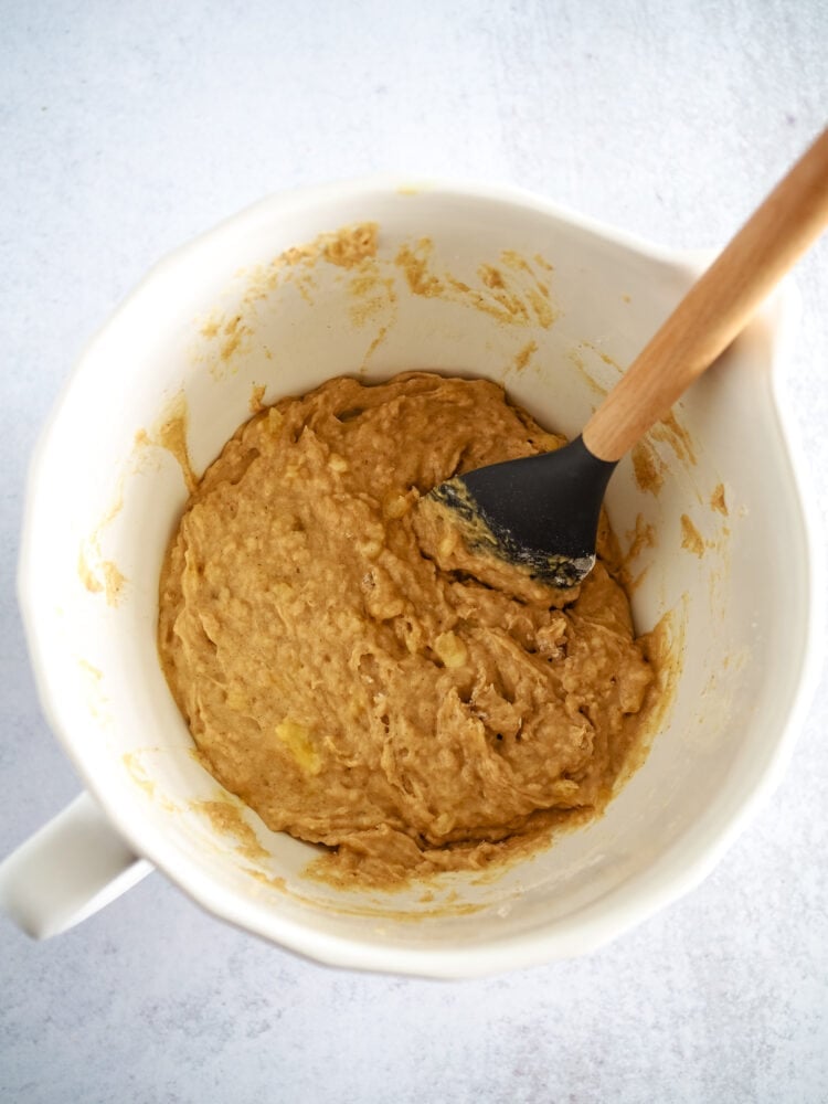 Ingredienst for banana nut muffins blended together with a grey silicone and wood spatula in a small white mixing bowl.