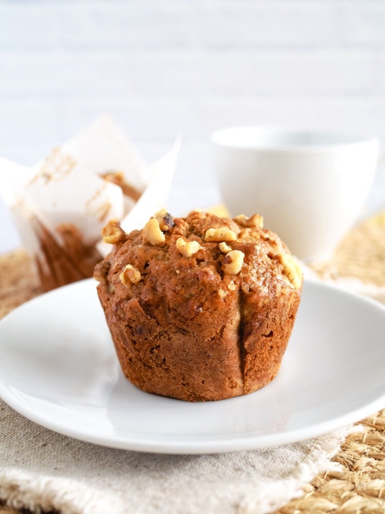 A single banana nut muffin on a white plate, resting on a natural linen napkin with a white coffee cup in the background.