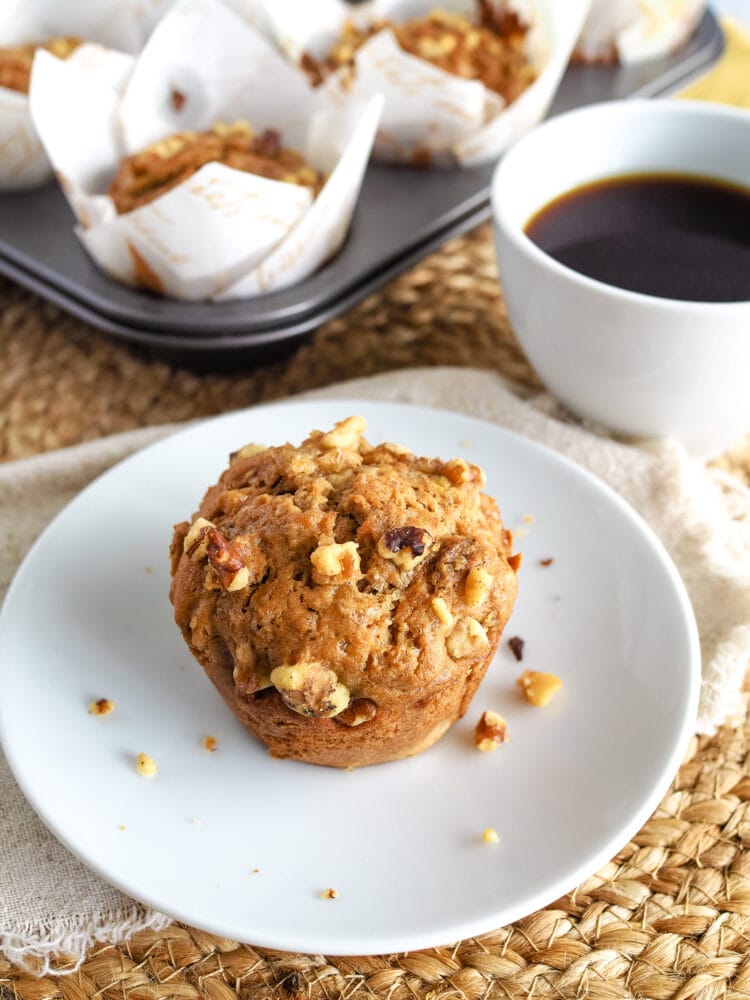 Banana nut muffin on a small white plate resting on a jute placemate. There's a coffee and a muffin tin full of banana nut muffins in the background.