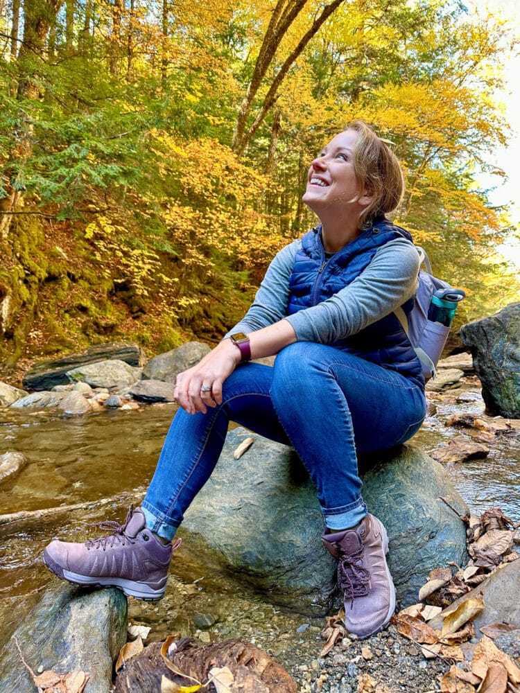 Rachelle sitting on a rock by a river looking up at autumn leaves.