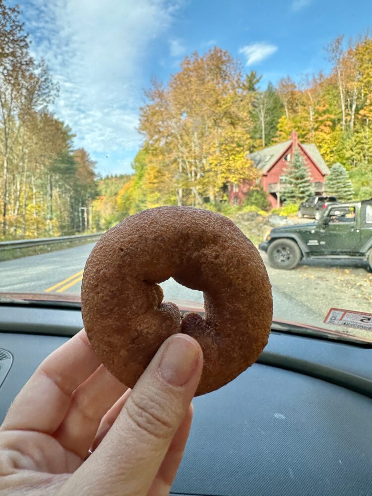 Rachelle holding up an apple cider donut while in the car driving around Stowe VT