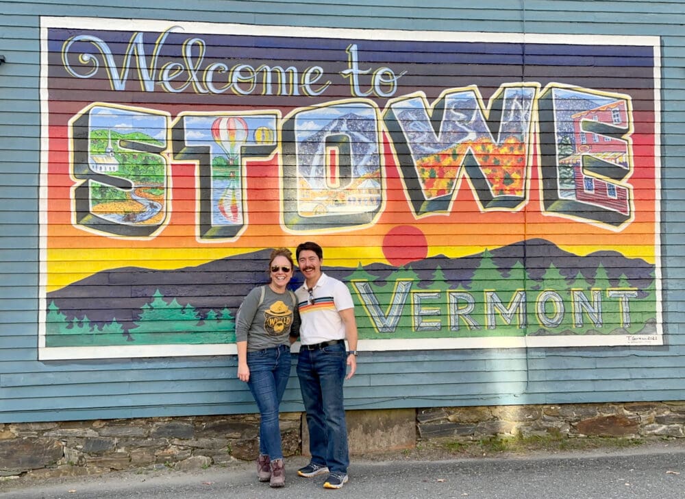 Rachelle and Pete standing in front of a mural painted to look like a postcard for Stowe, VT