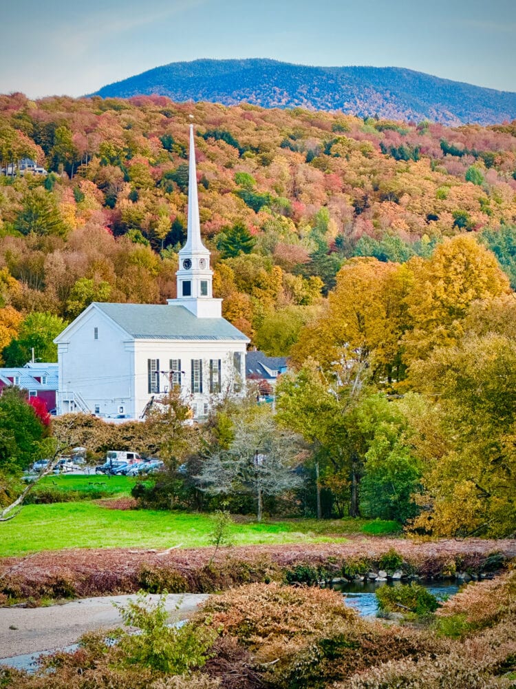 Photo of white church in Stowe, Vermont, with mountains in the background covered in autumn colored trees of orange, gold, red, and green.