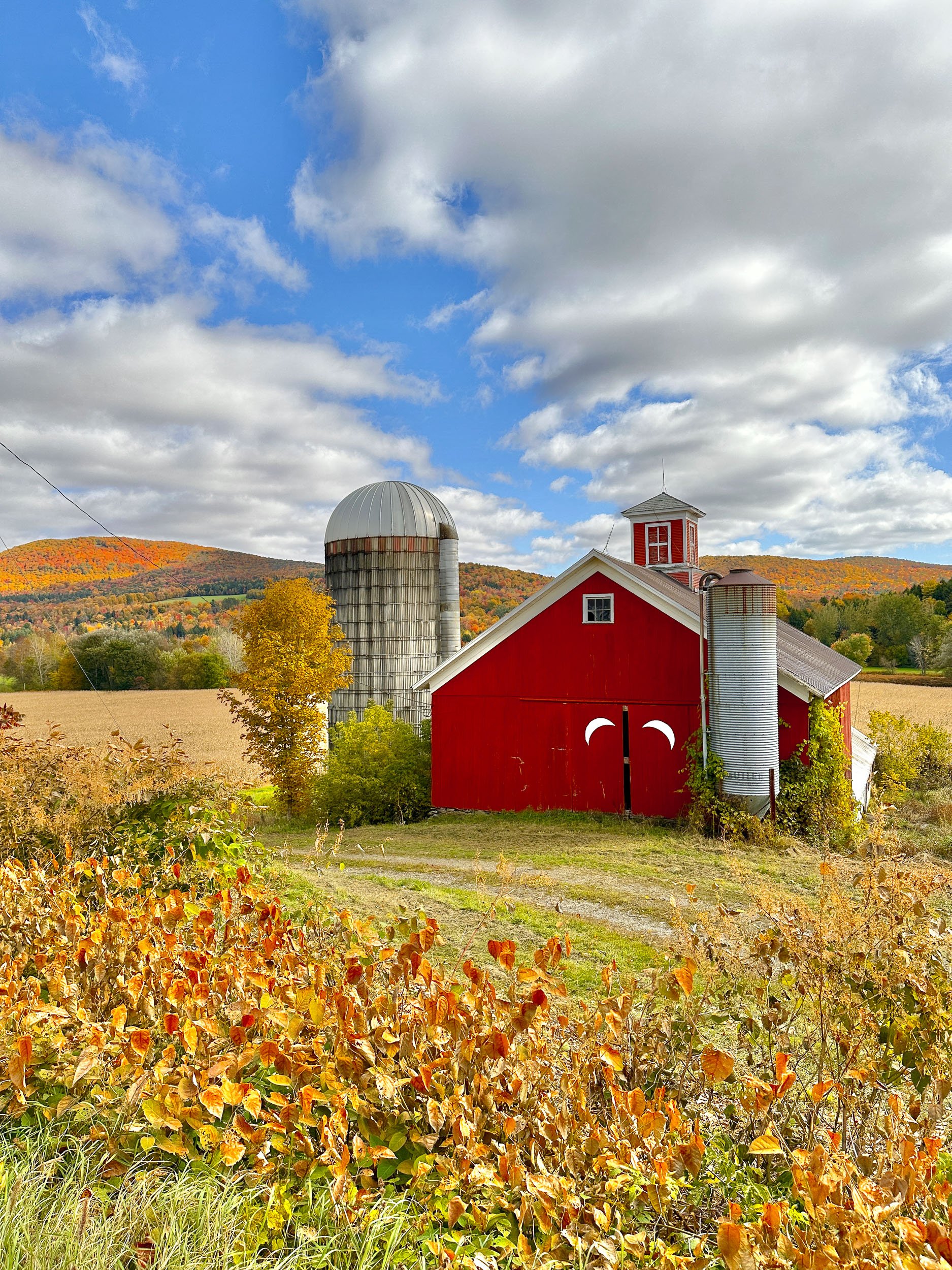 Photo of a red barn with wood silow sliver moons on the doors. The background is full of fall colored leaves on rolling hills. 