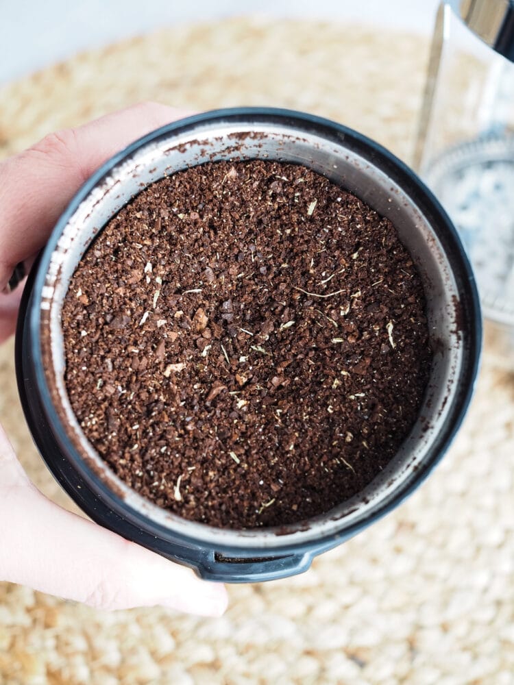 A mix of ground up light and dark coffee beans and cardamom pods in a coffee grinder.