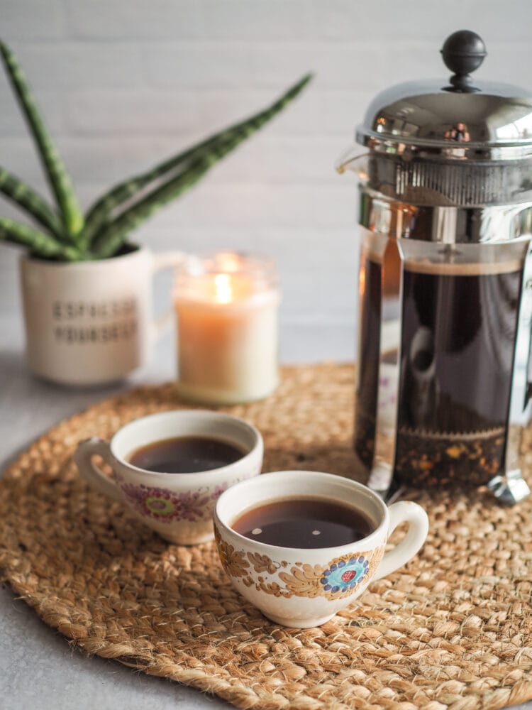 Two cups of small espresso sized cardamom coffee with a french press on a straw mat. In the background is a green snake plant and lit white candle.