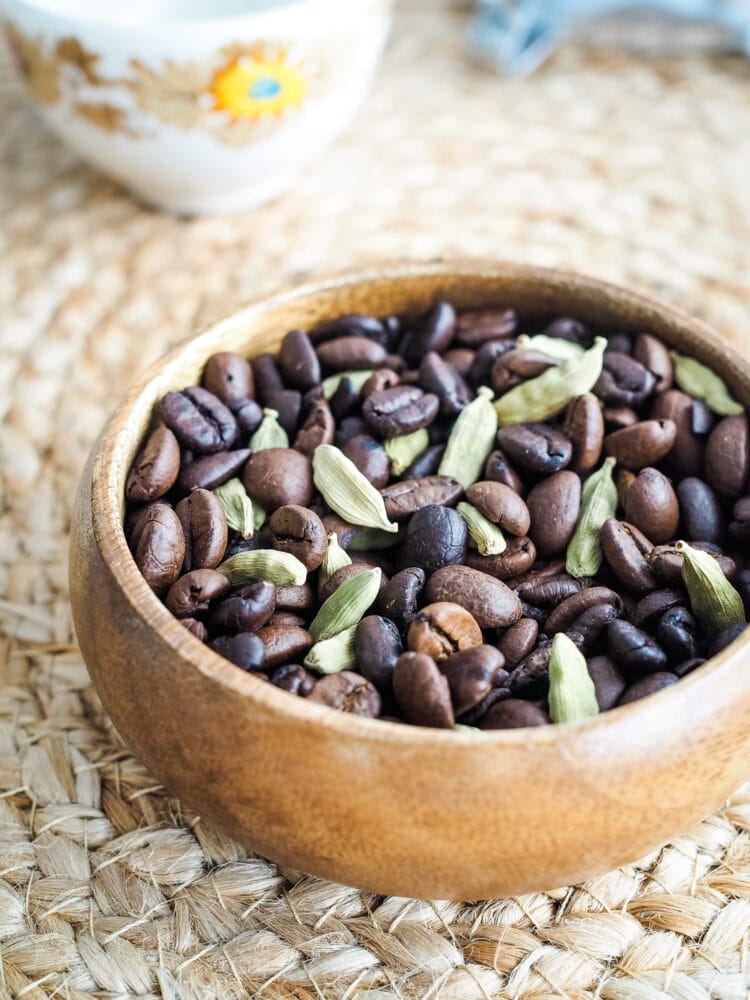 A mix of light and dark roast coffee beans with whole cardamom pods in a wood bowl