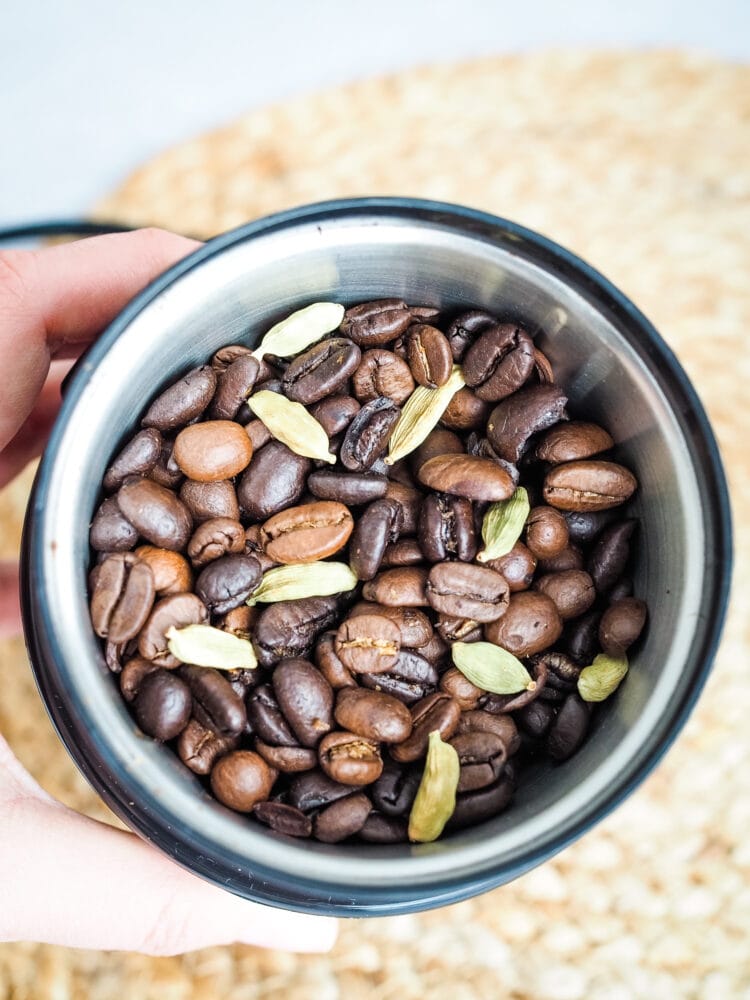 A mix of light and dark coffee beans and whole cardamom pods in a coffee grinder.