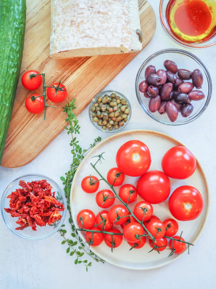 Overhead look at the ingredients needed to make dakos salad including crusty bread, cucumber, tomatoes, sun dried tomatoes, kalamata olives, capers, oregano, and olive oil and red wine vinegar.