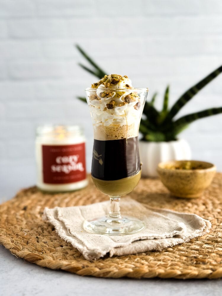 Pistachio macchiato with pistachio butter layered at the bottom of a glass, topped with espresso, whipped cream, and crushed pistachios.