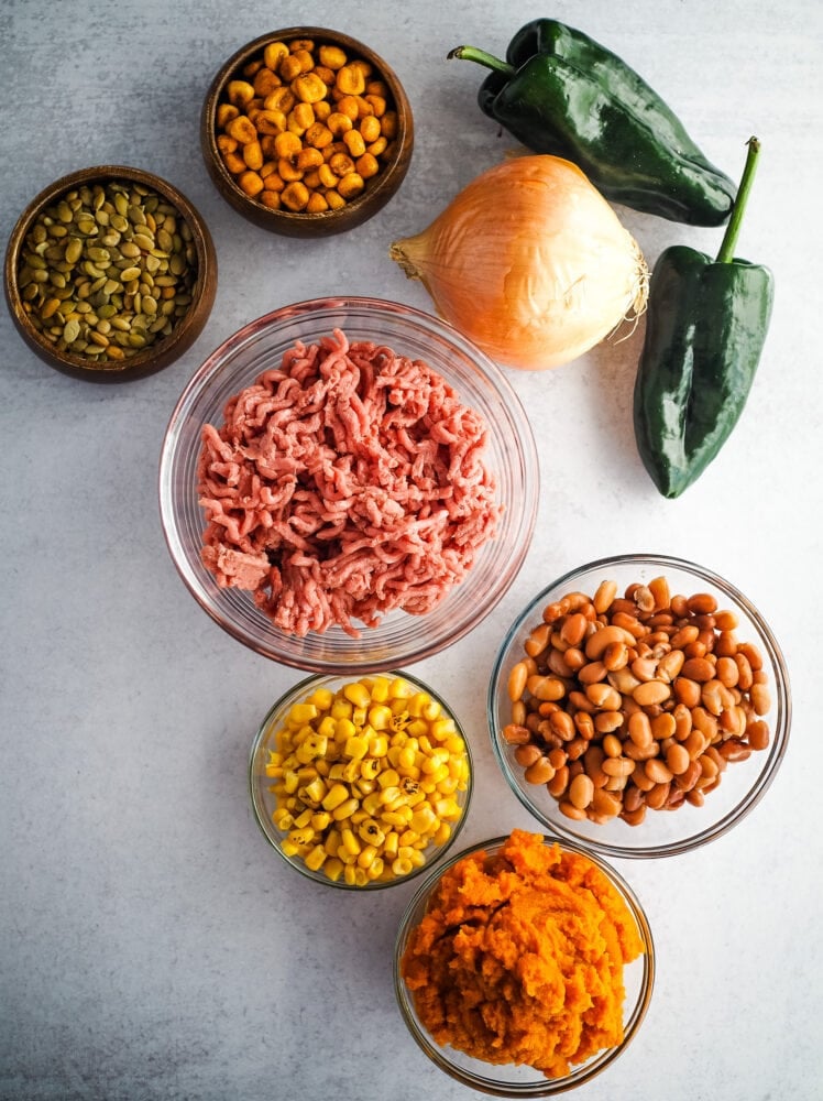 Ingredients and toppings for white bean turkey chili including ground turkey, white onion, poblano peppers, pinto beans, fire roasted corn, pumpkin puree, with pepitas and corn nuts for toppings.