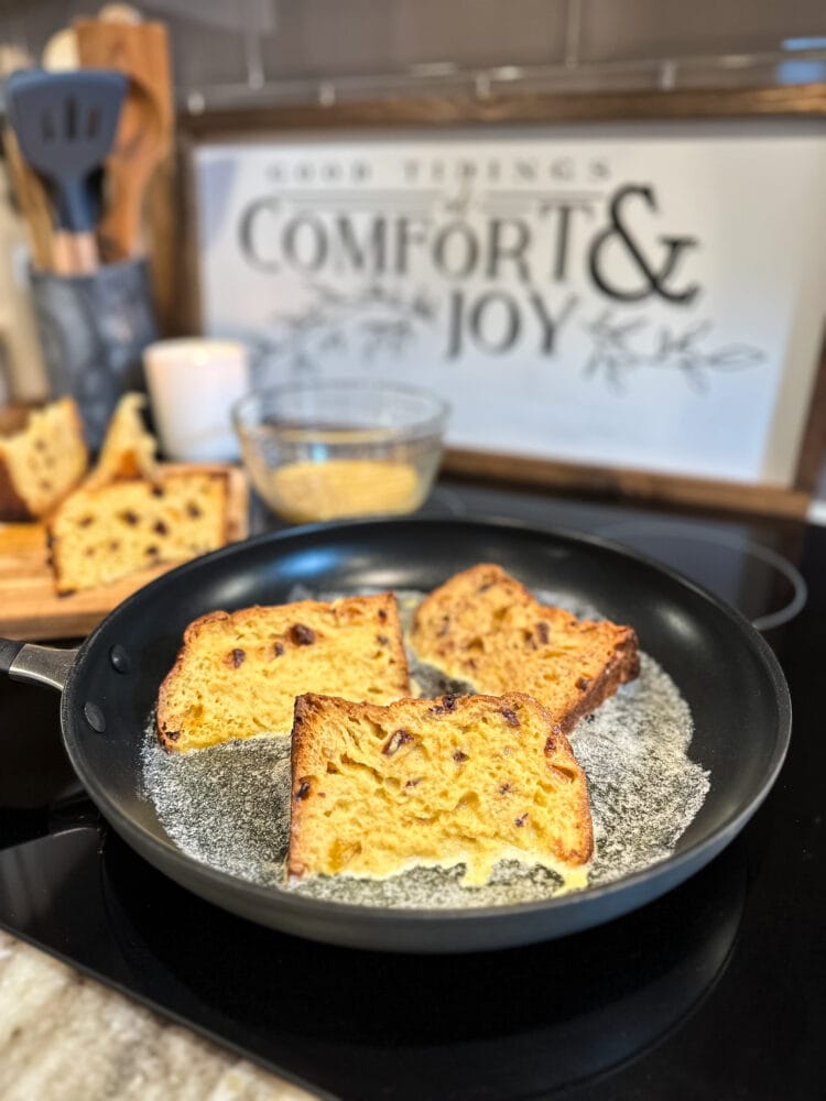 Four slices of panettone french toast in a skillet cooking. There's a black and white sign on the back of the stove that says "Good Tidings of Comfort and Joy" and a white candle burning, looking festive. 