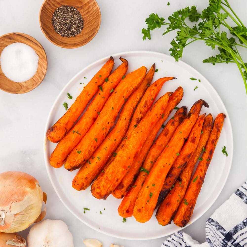 air fryer carrots, garnished with chopped parsley, on a white plate, with a white and blue striped kitchen towel and parsley on the side. There's also two small wood pinch bowls, salt in one, pepper in the other