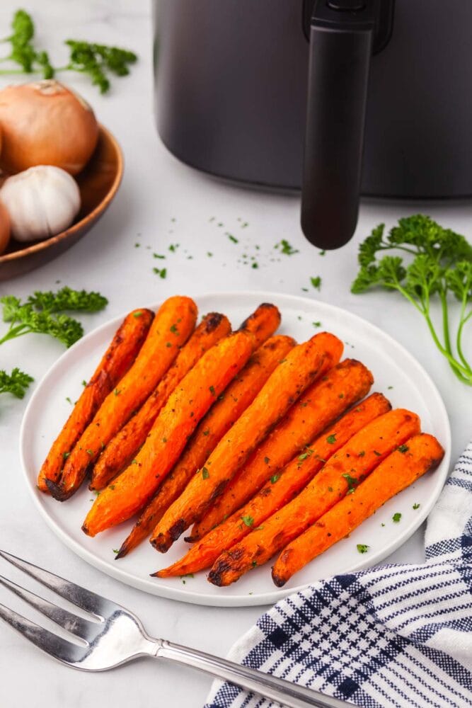 air fryer carrots, garnished with chopped parsley, on a white plate, with a white and blue striped kitchen towel and parsley on the side. There's also a wood bowl of onions and garlic, and a black air fryer in the background