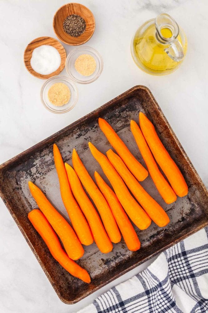 Whole peeled raw carrots on a sheet pan, with the seasoning spices and oil to the side and blue striped kitchen towel and parsley on the side.