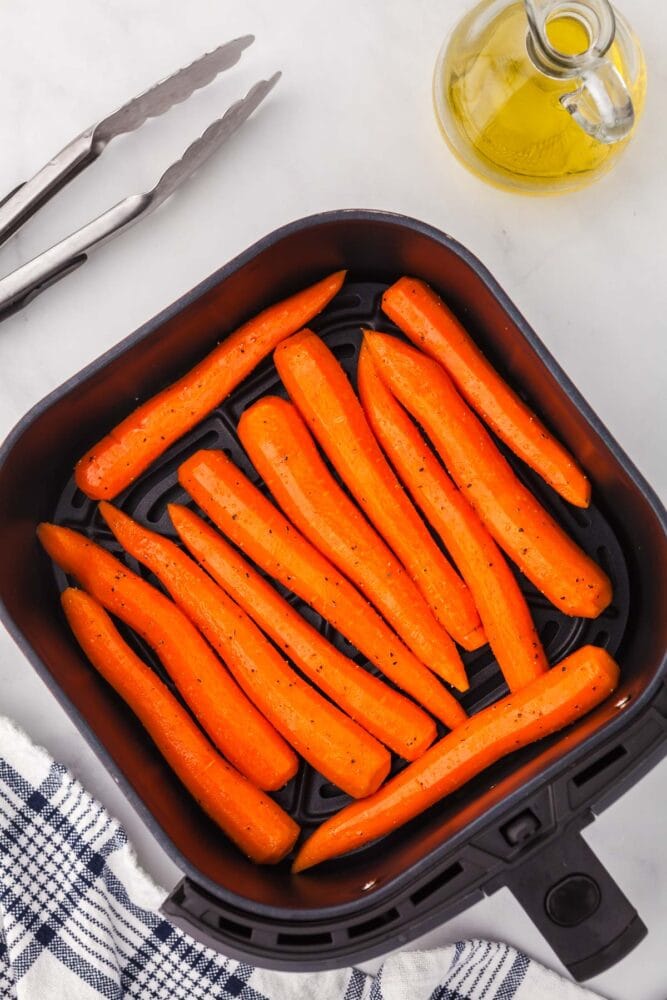 Whole, peepled, seasoned carrots in a square air fryer basket. There are tongs and a glass container of oil to the side as well as a blue and white striped kitchen towel.