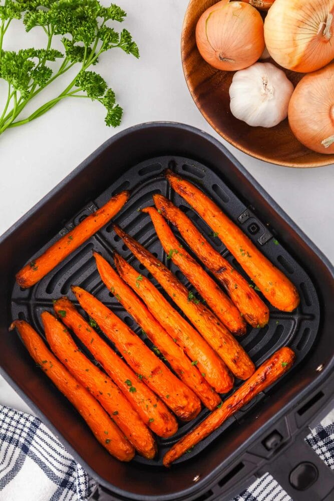 Air fried carrots in a square air fryer basket. They're slightly roasted and brown along the tips, and garnished with parsley. There's fresh parsely and a wood bowl of onions and garlic off to the side.