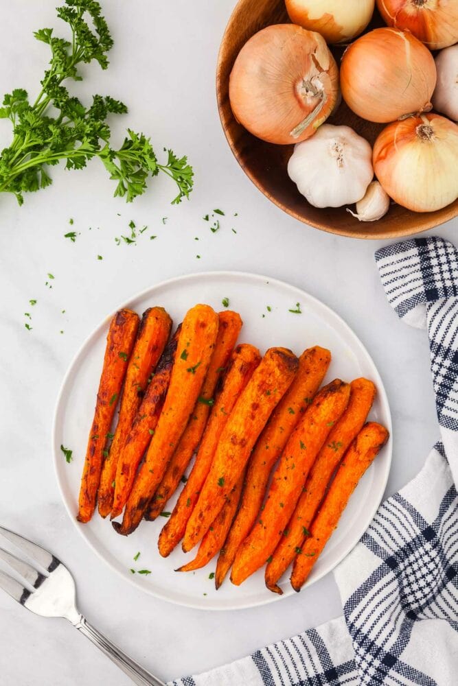 Air fryer carrots, garnished with chopped parsley, on a white plate, with a white and blue striped kitchen towel and parsley on the side. There's also a wood bowl of onions and garlic.