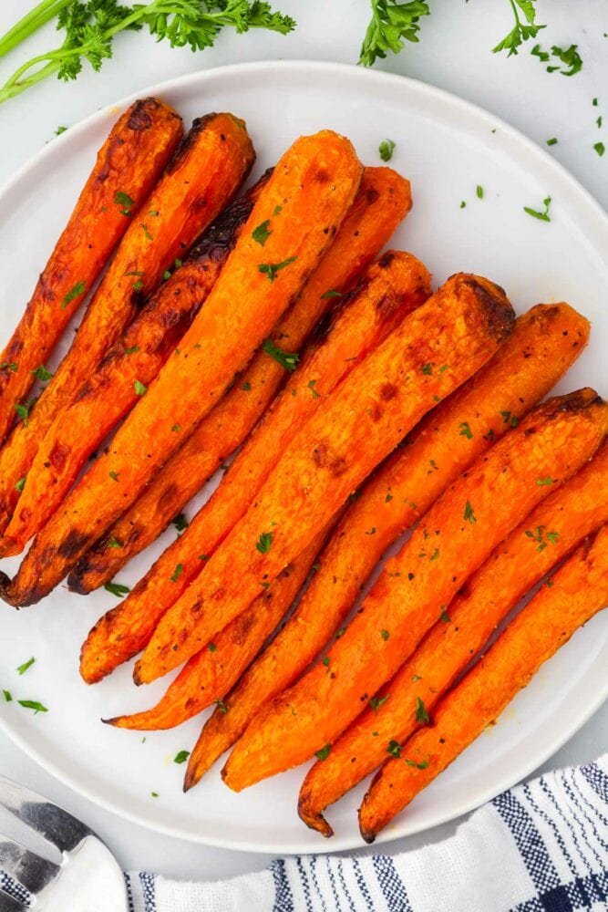 air fryer carrots, garnished with chopped parsley, on a white plate, with a white and blue striped kitchen towel and parsley on the side.