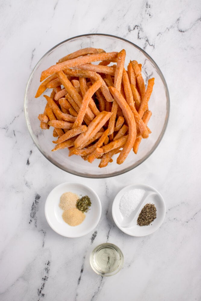 clear glass bowl with frozen sweet potato fries, two small white bowls filled with seasonings and spices, one small clear glass bowl with about a tablespoon of oil